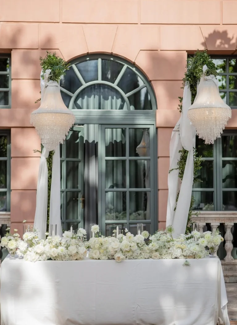 Crystal chandeliers above a table of white flowers before the hotel's pink façade