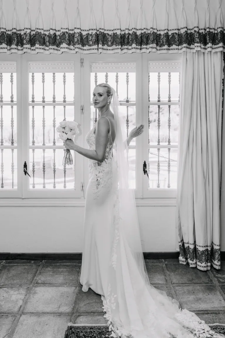 The bride, with a sleek updo, veil and white roses, looks back by a barred window