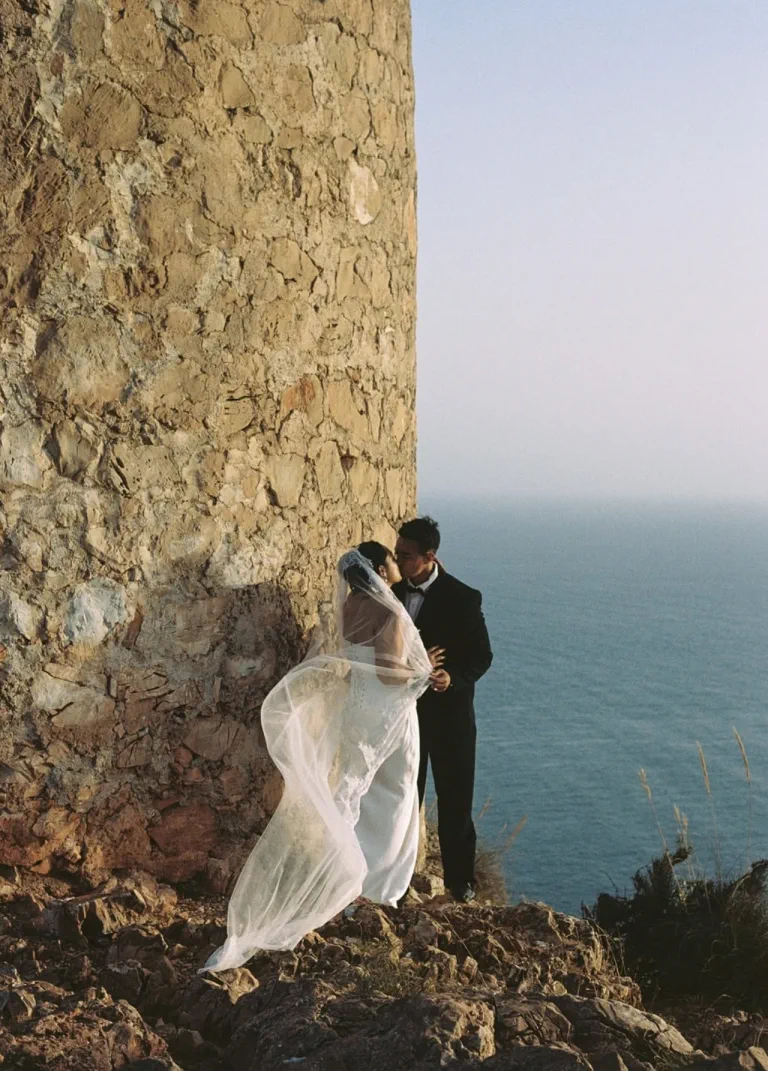 The couple kiss beside a stone tower, the veil in the wind and the sea behind them