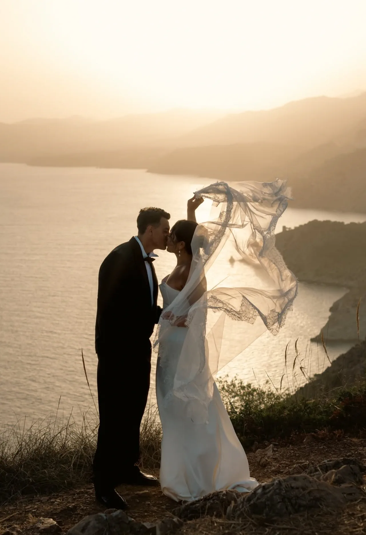 The couple kiss on the clifftop, her veil in the wind and the bay at sunrise behind them