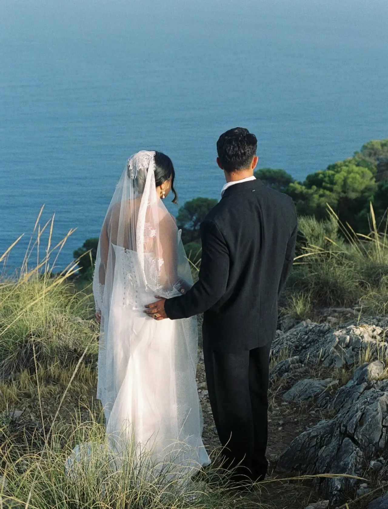 The couple from behind looking out to sea, the long veil falling over the bride’s updo
