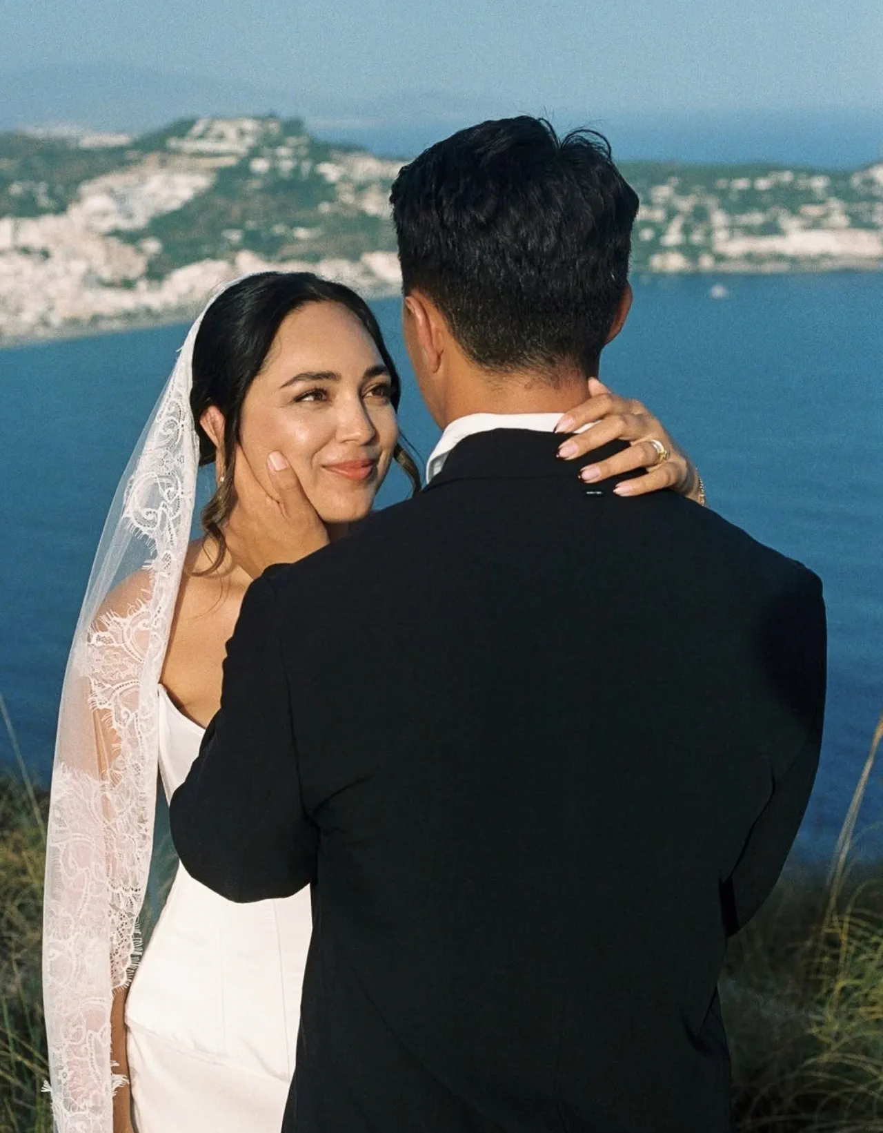 Bride with a low updo, a long lace-edged veil and luminous makeup, embracing her husband above the sea