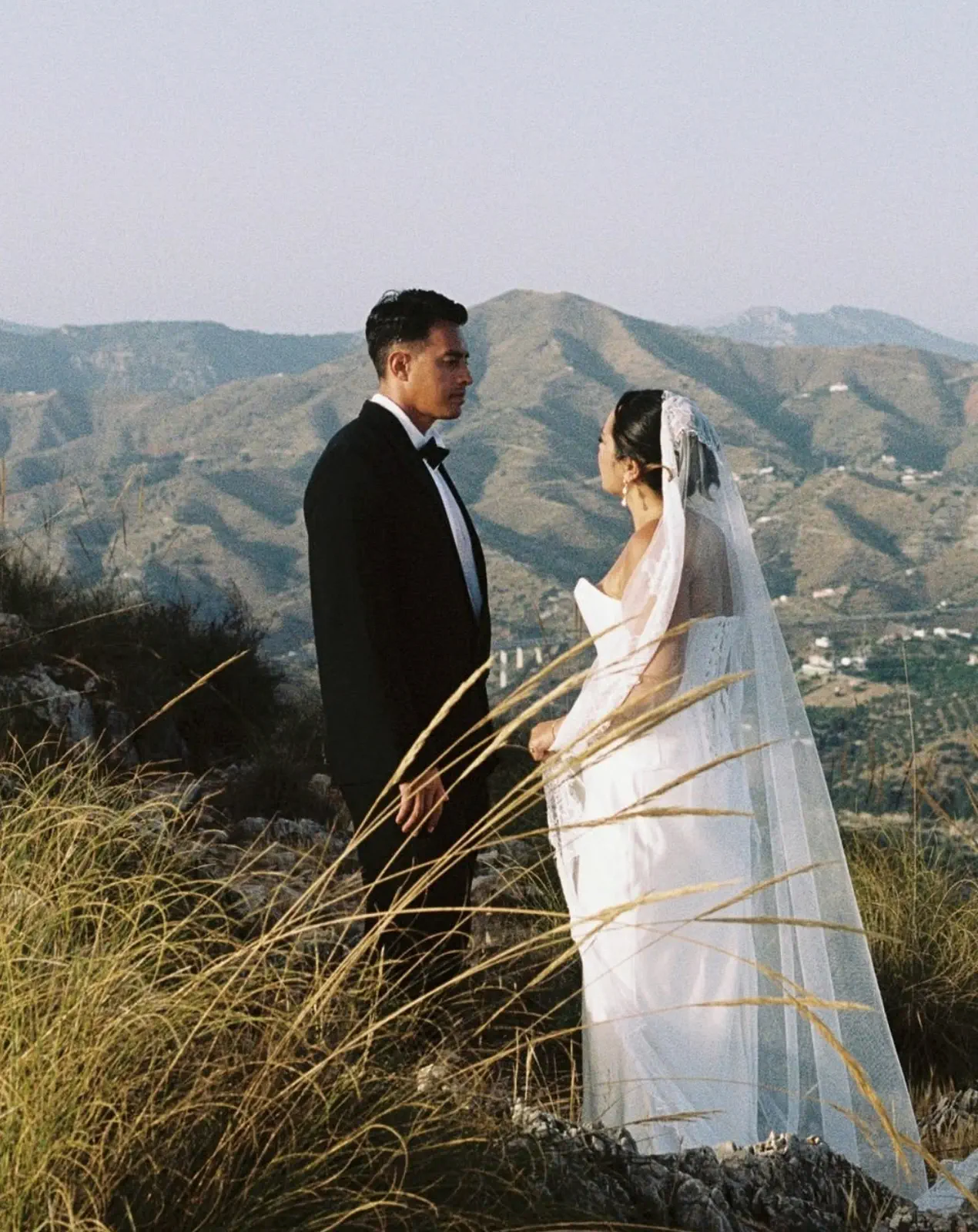 The couple look at each other among tall grass, with the mountains and the sea behind