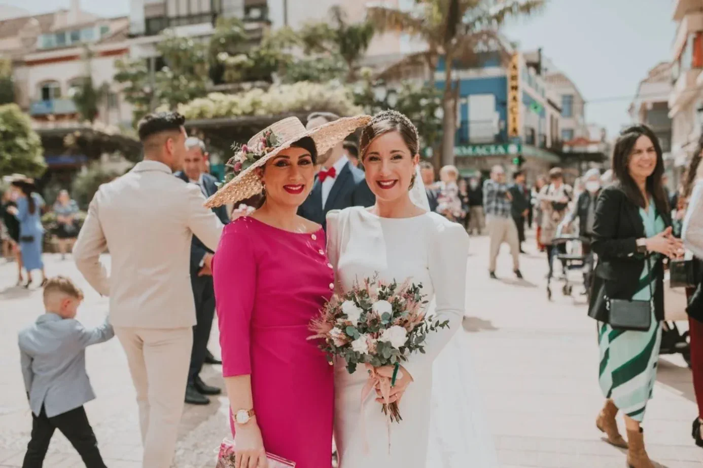The bride, holding her bouquet, beside a guest in a wide-brimmed hat in the square of the church of El Rosario, Fuengirola