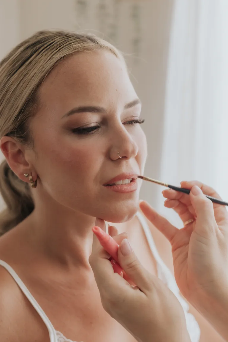 Close-up of the bride with her eyes closed as her lips are lined, wearing soft glam makeup with luminous skin