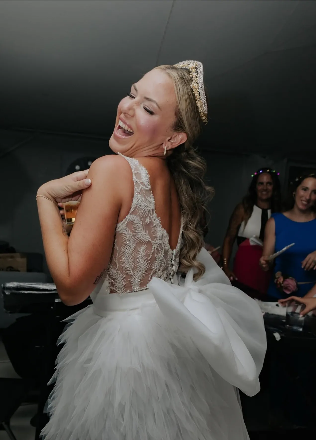 The bride laughs, turning away mid-party, wearing a gold tiara and a white feathered dress