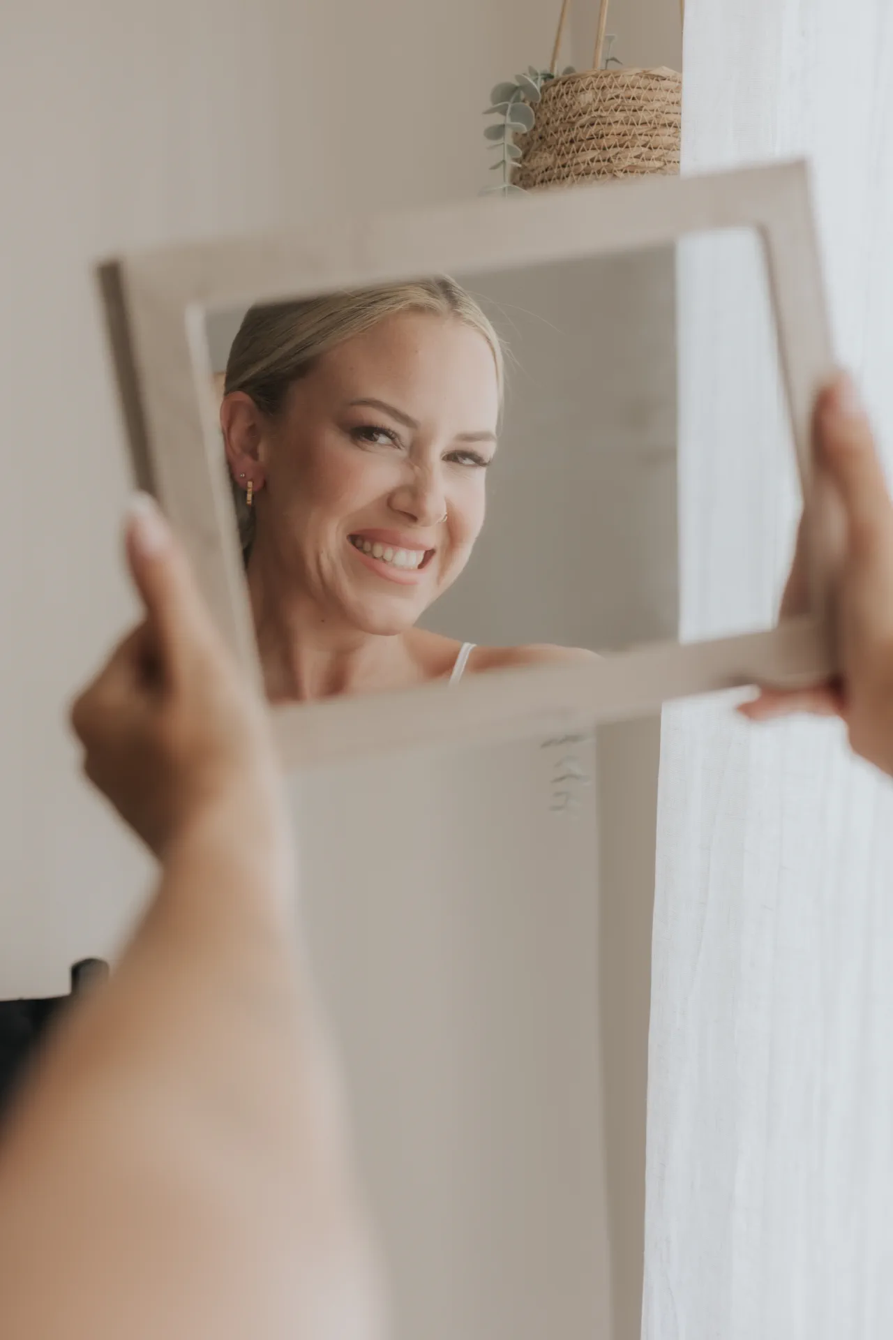 Bride smiling into a hand mirror with luminous soft glam makeup and pink blush