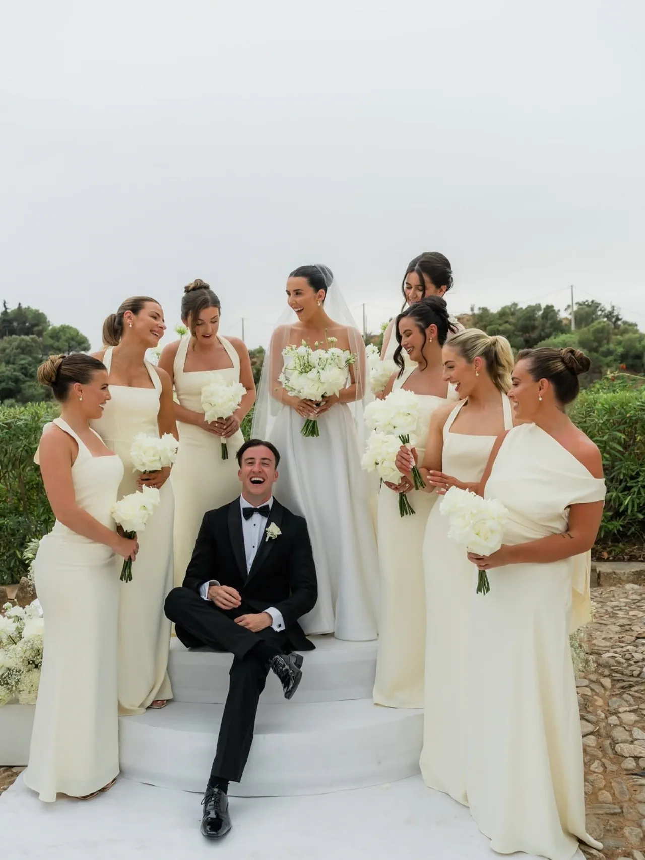 The bridesmaids, with sleek updos, laugh around the groom sitting on white steps