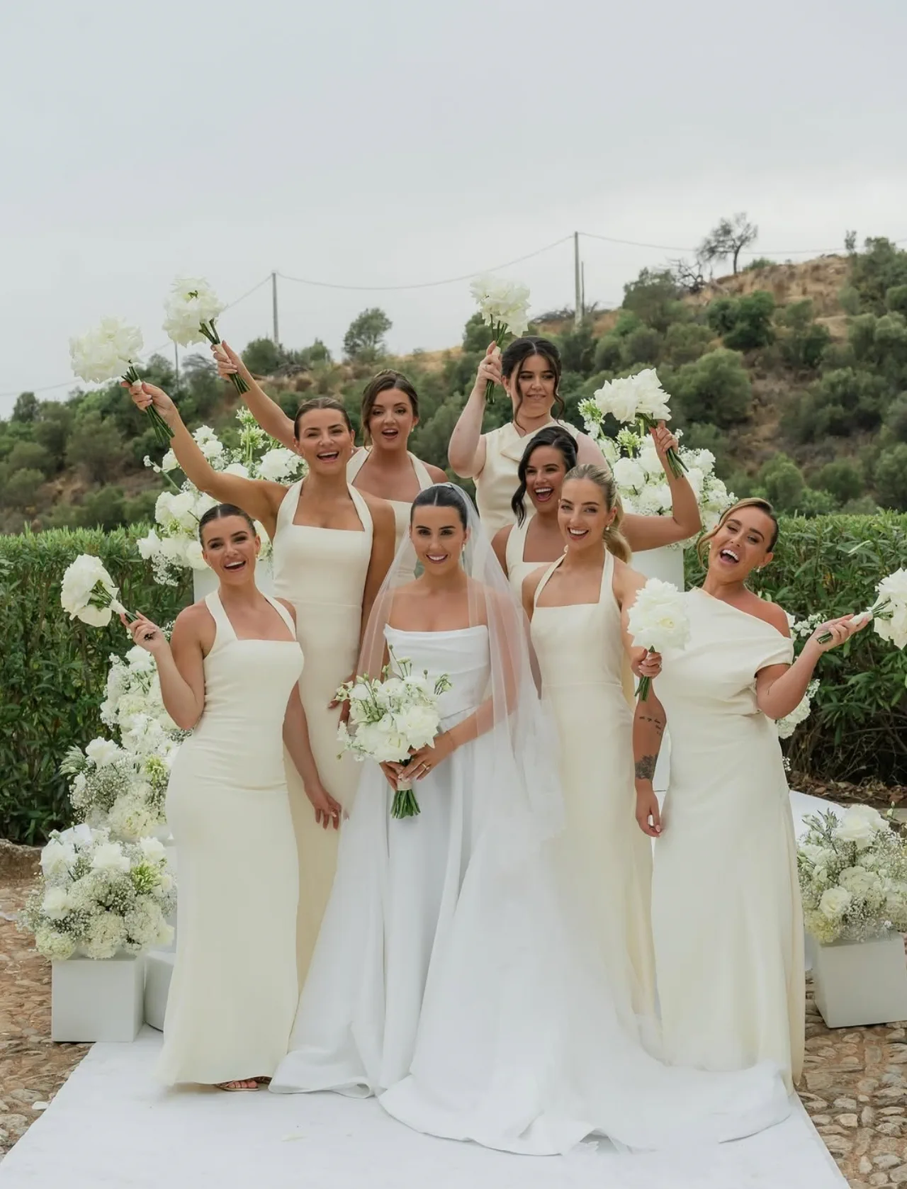 The bridesmaids, in cream dresses with sleek hairstyles, raise their white bouquets around the bride