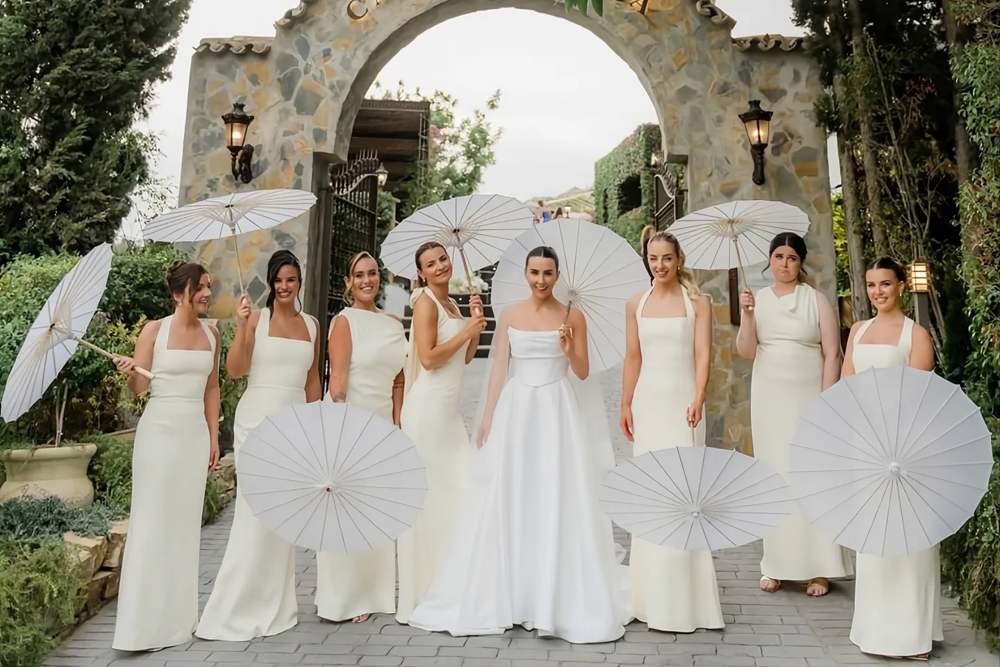 The bride and her bridesmaids with white parasols under the stone arch of Casa de la Era