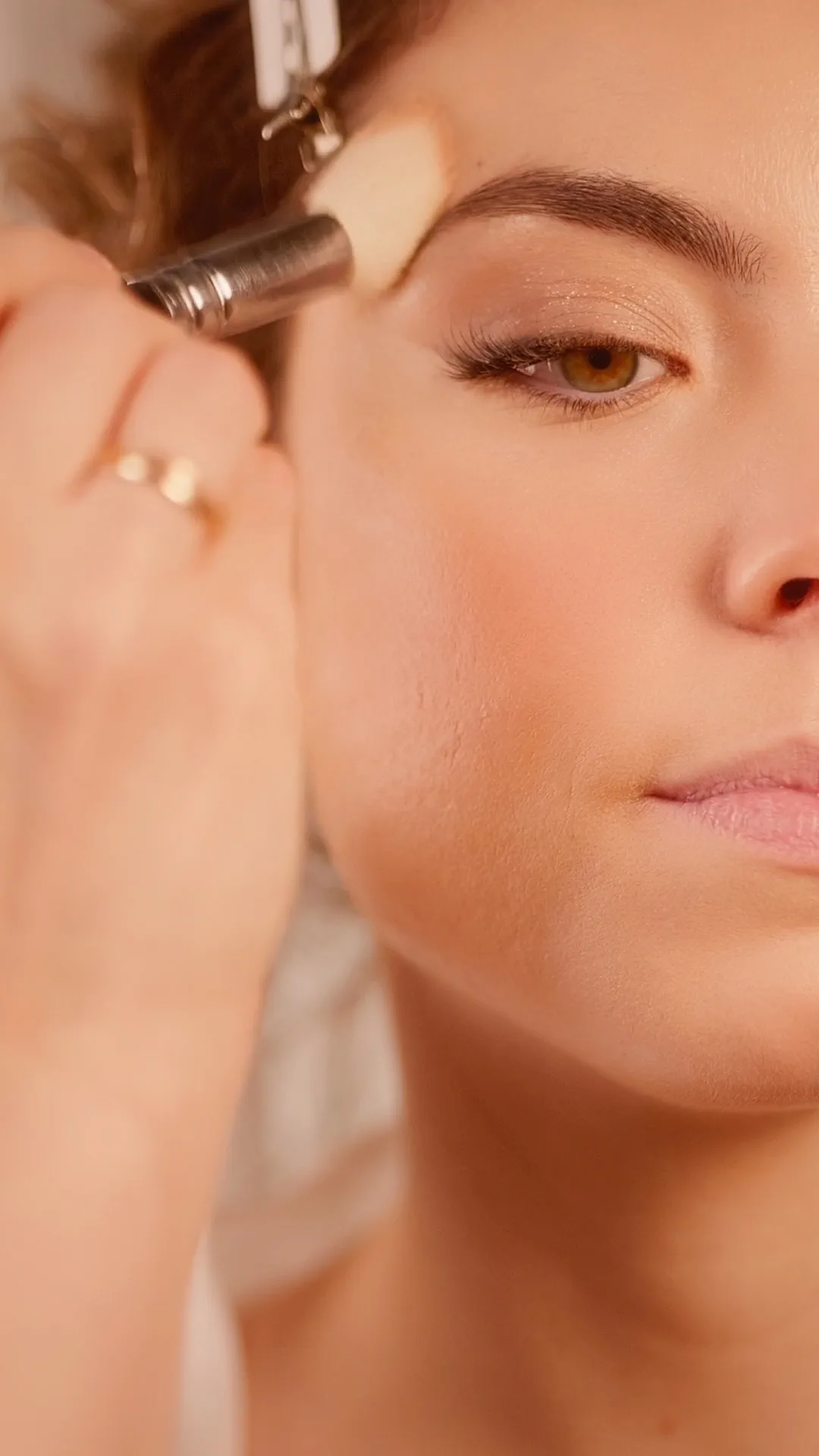 Bride during her makeup, with the brush on her brow and the texture of her skin still visible